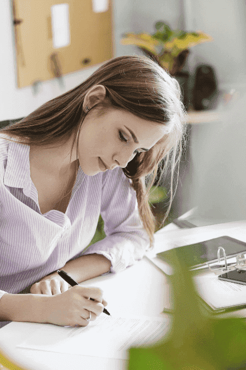 Woman writing at desk, pen in hand, concentrating on paper in front of her.